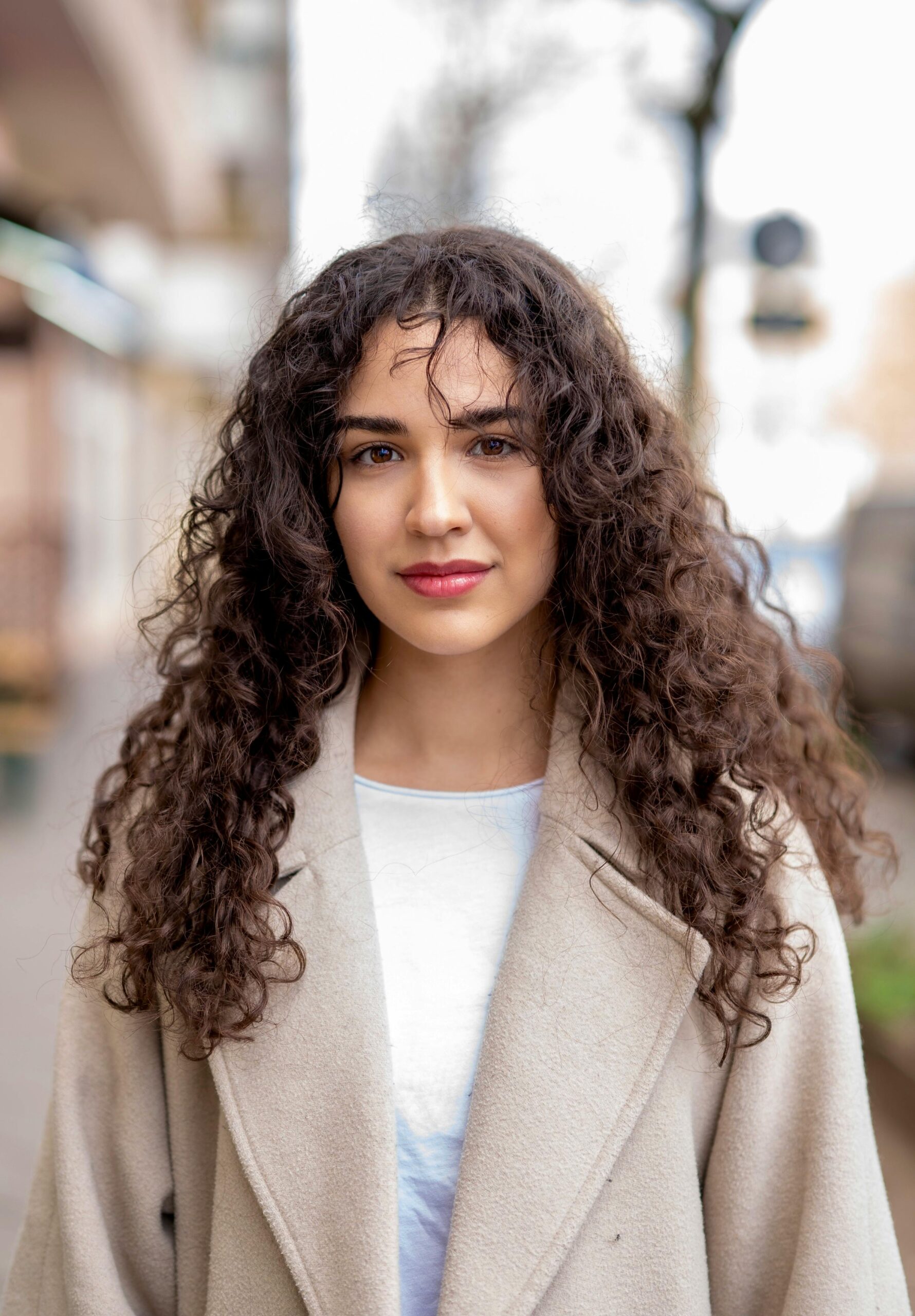 A young woman with curly hair and red lips, wearing a coat, posing outdoors in Berlin.
